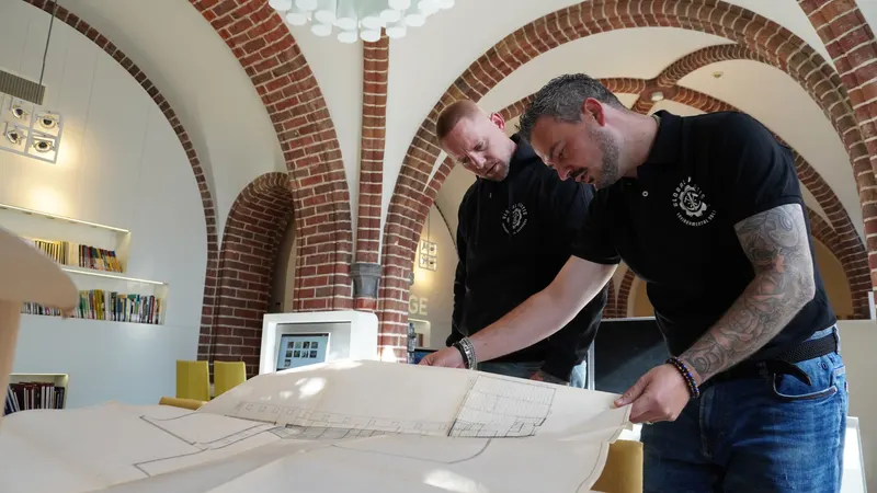 Twee medewerkers in bedrijfskleding bekijken samen een bouwtekening in het kantoor